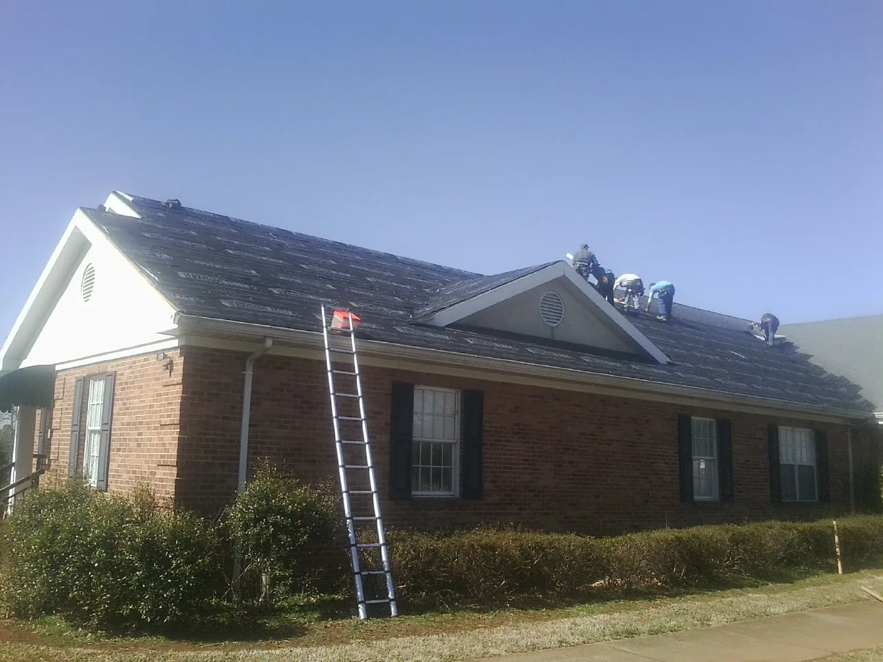 Crew tearing shingles off a brick ranch roof with a ladder set — Metal Roof Repair work by our crew in Lake Como
