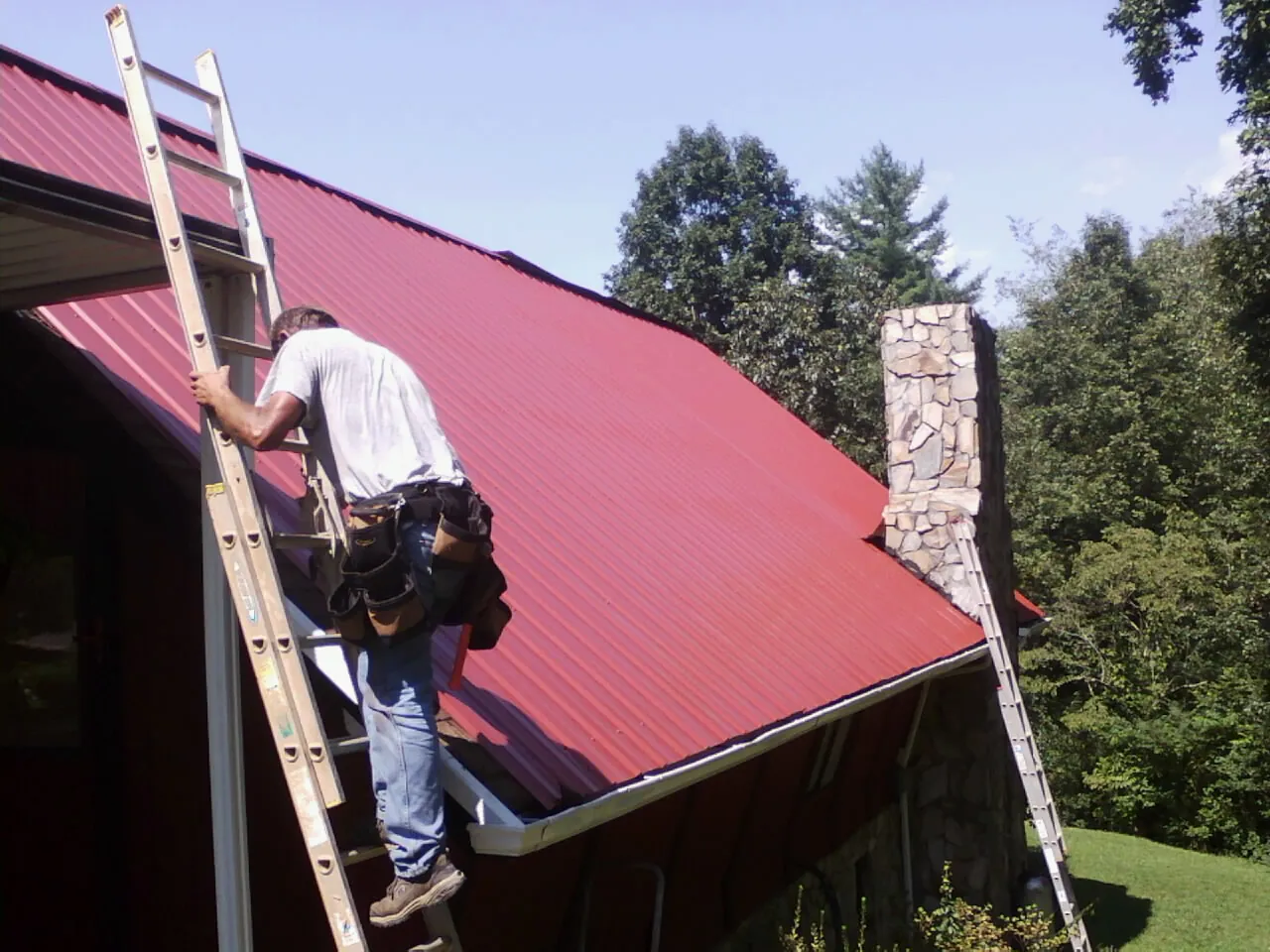 Roofer on a ladder beside a red metal roof and stone chimney — Soffit & Fascia Repair by Roofing Brothers in Lake Como